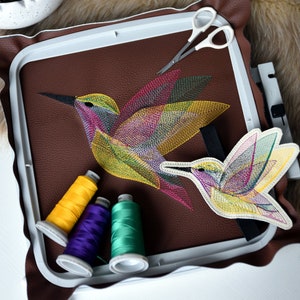 Può includere: Due colibrì ricamati colorati su uno sfondo di cuoio marrone in un telaio da ricamo. Ci sono tre bobine di filo in giallo, viola e verde.