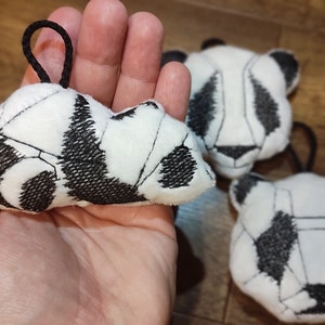May include: Three black and white panda plush toys. The panda on the left is lying down and has a loop for hanging. The other two pandas are standing up and have a more traditional panda shape.