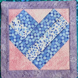 May include: A handmade heart-shaped quilted coaster with a pink background and a blue and white patterned heart in the center. The coaster is surrounded by a purple border with a decorative stitch.