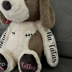 Pode incluir: Um c&atilde;o de pel&uacute;cia branco e marrom com detalhes bordados. O c&atilde;o tem a l&iacute;ngua rosa para fora e as palavras "My Tatas" e "Kathy" s&atilde;o bordadas nas patas. As palavras "ukunna" e "2/23" s&atilde;o bordadas no corpo.