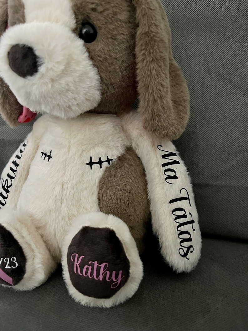 Pode incluir: Um c&atilde;o de pel&uacute;cia branco e marrom com detalhes bordados. O c&atilde;o tem a l&iacute;ngua rosa para fora e as palavras "My Tatas" e "Kathy" s&atilde;o bordadas nas patas. As palavras "ukunna" e "2/23" s&atilde;o bordadas no corpo.