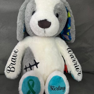 May include: A white and gray plush dog with a blue and green ribbon on its paw. The dog has the words "Brave" and "Warrior" embroidered on its arms and the name "Sterling" on its other paw.