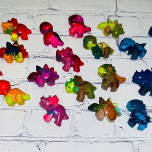 May include: A collection of 20 colourful dinosaur-shaped crayons. The crayons are made of wax and have a variety of colours, including red, orange, yellow, green, blue, and purple.