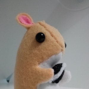 May include: A small, beige and white plush toy hamster with black eyes and pink ears. The hamster is holding a black and white striped object in its paws.