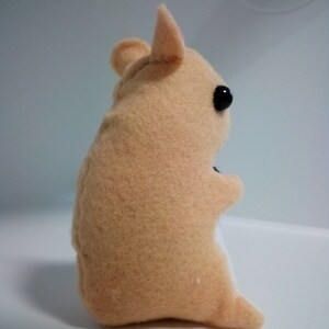 May include: A small, beige plush hamster toy with black eyes and a pink nose. The toy is sitting on a white surface.