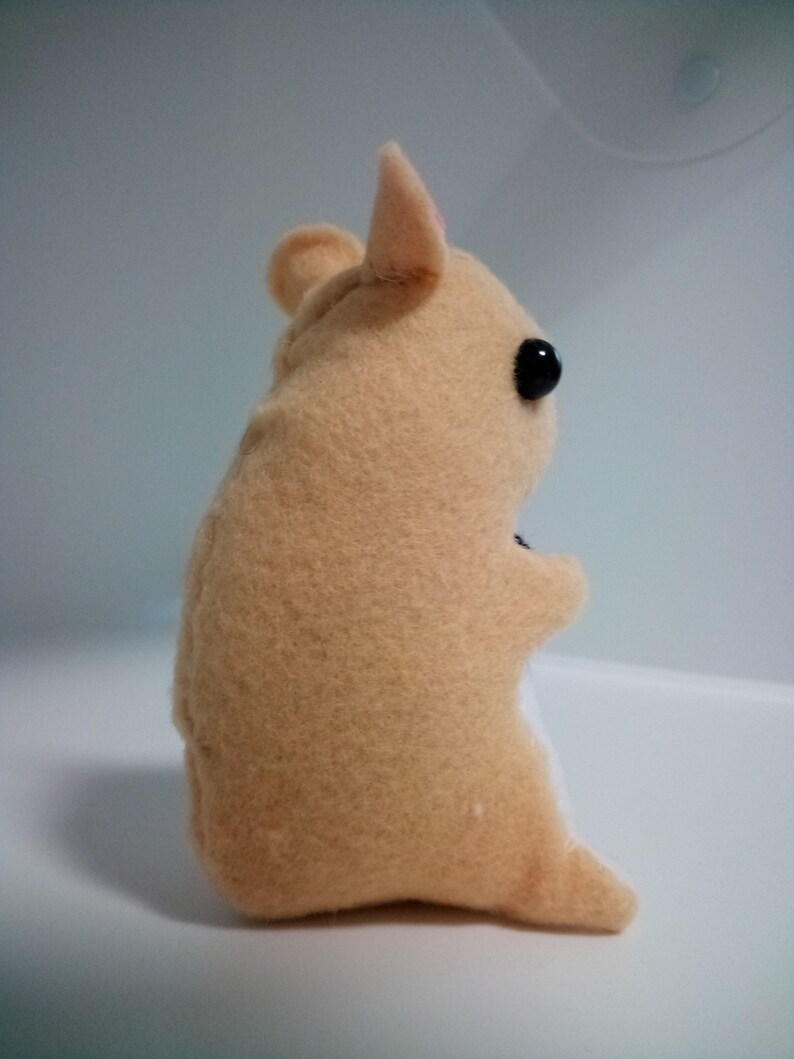 May include: A small, beige plush hamster toy with black eyes and a pink nose. The toy is sitting on a white surface.