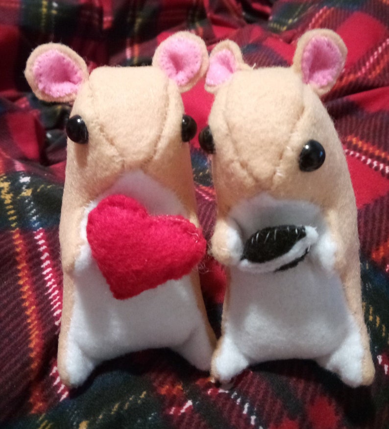 May include: Two plush brown and white hamster toys. One hamster is holding a red heart and the other is holding a black and white cookie.