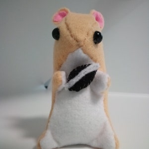 May include: A small, tan plush hamster toy with pink ears and black eyes. The hamster is holding a black and white cookie in its paws.