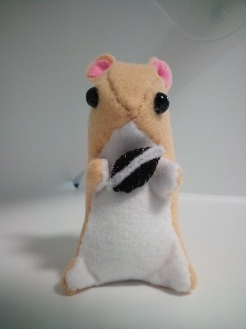 May include: A small, tan plush hamster toy with pink ears and black eyes. The hamster is holding a black and white cookie in its paws.