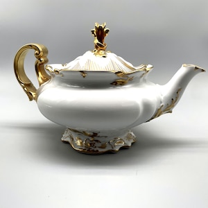 May include: A white porcelain teapot with gold accents. The teapot features a curved handle, a decorative lid with a gold finial, and gold trim along the edges. The spout and base also have gold detailing.