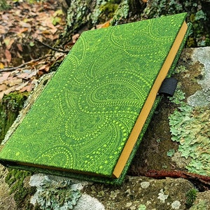 Può includere: Un diario verde con un motivo paisley e bordi dorati, appoggiato su una roccia ricoperta di muschio in un ambiente boschivo.