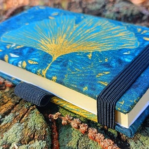 May include: A blue and gold patterned notebook with a black elastic closure. The notebook is closed and lying on a textured surface.