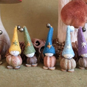 May include: A group of charming gnome ornaments, each with a felt hat and wooden body. The hats are in various colours, such as red, yellow, blue, and purple, and topped with a bell. The gnomes have painted faces and are displayed on a green surface.