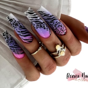 May include: Long, square-shaped artificial nails with a gradient design. The nails transition from pink to purple, featuring black zebra stripes and leopard spots. Gold rings adorn the fingers. The Decora Nails logo is visible.