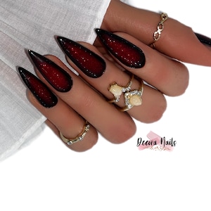 Könnte beinhalten: Nahaufnahme einer Hand mit langen, stilettoförmigen Nägeln, die mit einem schwarzen und roten Ombre-Design lackiert sind. Die Nägel sind mit Glitzer verziert. Die Hand trägt mehrere goldene Ringe, darunter einen mit einem weißen Stein.