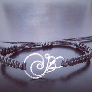 May include: A black macramé bracelet with a silver-coloured wire design. The design features a stylised letter "B" with a spiral element. The bracelet has adjustable sliding knots for a custom fit.