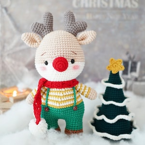 May include: A crocheted reindeer toy with a red nose, a red and white striped scarf, and green pants. The reindeer is standing next to a crocheted green Christmas tree with a white star on top.