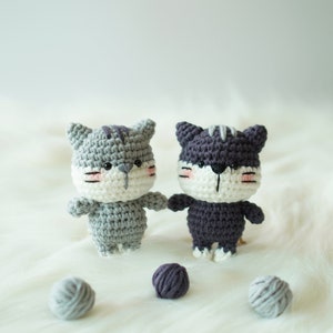 May include: Two crocheted cat toys, one grey and one purple, with white faces and pink cheeks. The toys are sitting on a white fluffy surface with balls of yarn.