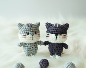 May include: Two crocheted cat toys, one gray and one purple, with white faces and pink cheeks. The toys are sitting on a white fluffy surface with balls of yarn.