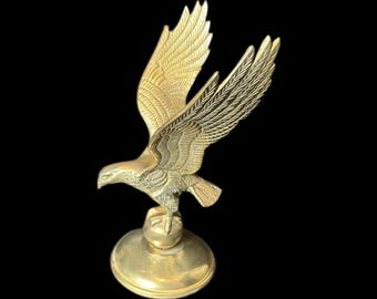 May include: A gold-colored metal eagle figurine with its wings spread, perched on a round base. The eagle is in flight, with its head turned to the side.