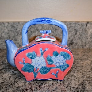 May include: A ceramic teapot featuring a coral-colored body with a blue and white floral pattern. The teapot has a blue arched handle, a small spout, and a matching lid with a knob. The design includes green leaves and white blossoms.
