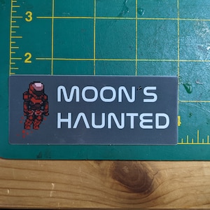 Può includere: Un adesivo grigio con testo bianco che recita "MOON'S HAUNTED". Un'immagine pixelata di un astronauta rosso si trova sul lato sinistro dell'adesivo.