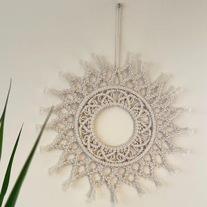 Op de afbeelding: Een witte macramé wandhanger in de vorm van een zon met een cirkelvormig ontwerp in het midden. De hanger heeft een franje rond de rand.
