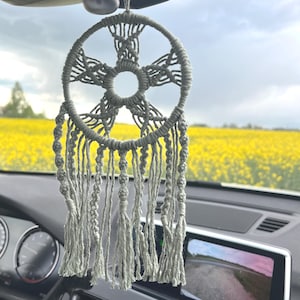 Könnte beinhalten: Ein Traumfänger aus salbeigrünem Makramee, der am Rückspiegel eines Autos hängt. Das kreisförmige Design weist komplizierte Knoten und lange, herabhängende Fransen auf. Der Hintergrund zeigt ein Feld mit gelben Blumen unter einem bewölkten Himmel.