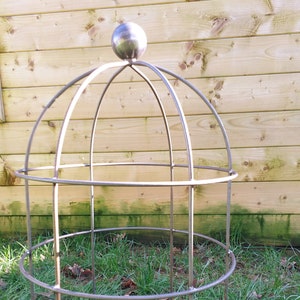May include: A metal plant cage with a rounded top and a silver ball finial. The cage is made of thin metal rods and has two circular rings. The cage is standing on a bed of green grass.