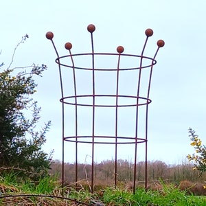 May include: A rusty metal plant support with a round top and six rounded points. The support has three horizontal rings.
