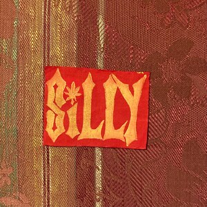 May include: A red fabric patch with the word "SILLY" written in gold lettering. The letters are stylized and have a jagged, almost gothic, appearance.