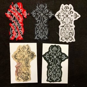 May include: Five fabric patches with a decorative, ornate design. The patches are in various colors, including black, white, red, and beige. The design is a stylized cross with intricate details.