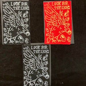Puede incluir: Tres parches de tela negra con texto e imágenes bordadas en blanco. El texto en cada parche dice "Look for the signs". Cada parche presenta una imagen estilizada de un pájaro con las alas extendidas y múltiples ojos que lo rodean.