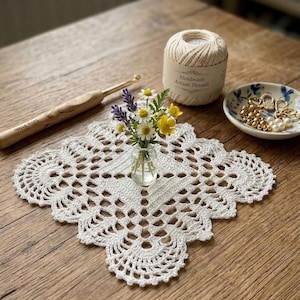 May include: A white square crocheted doily with a scalloped edge, a small glass vase containing yellow and purple flowers, a wooden crochet hook, a ball of cream-colored yarn labeled Handmade Artisan Threads, and a small bowl of gold beads.