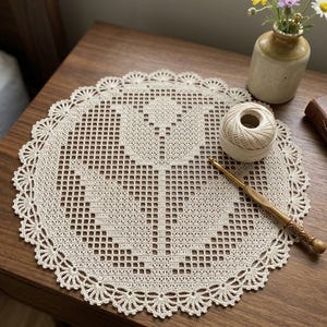 Puede incluir: Un tapete de ganchillo redondo de color blanco roto con un motivo central de tulipán creado con un patrón de rejilla. La pieza circular tiene un borde festoneado decorativo con detalles en forma de abanico, hecho de hilo fino y claro.