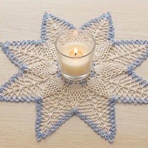 May include: A star-shaped crochet doily featuring cream-colored thread with light blue accents along the edges. A clear glass votive candle holder containing a lit white candle sits in the center. A small, metallic gold-toned thimble is nearby.