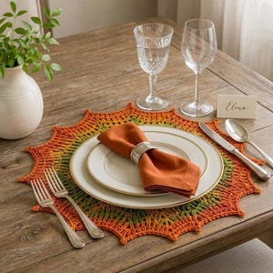 May include: A round, star-shaped crochet placemat featuring a gradient of orange, yellow, and green yarn. Centered on the placemat is a white dinner plate with a gold rim, topped with a folded orange cloth napkin held by a silver napkin ring.