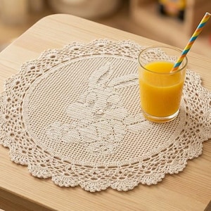 Può includere: Un centrino rotondo all'uncinetto color crema con un motivo centrale a forma di coniglio e un bordo in pizzo smerlato. Un bicchiere trasparente pieno di succo d'arancia con una cannuccia a righe colorate poggia sul centrino, sopra un tavolo in legno chiaro.