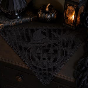 Puede incluir: Un tapete triangular de ganchillo negro con un patrón detallado de filet que representa una calabaza con sombrero de bruja, acentuado con un motivo de murciélago. Los bordes están rematados con un delicado festoneado decorativo.