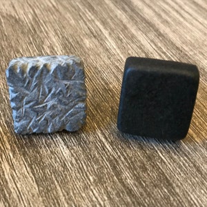 May include: Two square-shaped objects. One is a textured, gray stone with etched lines. The other is a smooth, black square. Both are displayed on a wooden surface. These could be decorative elements or components of a larger design.
