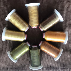 Puede incluir: Vista cenital de ocho carretes de hilo metálico en tonos dorados, bronce y verde. Cada carrete está envuelto en plástico transparente y tiene una base blanca con un anillo rojo. Los hilos están dispuestos en un patrón circular, mostrando una variedad de colores.