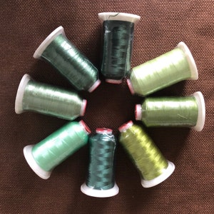 Puede incluir: Vista cenital de ocho carretes de hilo de bordar verde dispuestos en un patrón circular. Los colores del hilo varían de verde claro a oscuro. Cada carrete tiene una base blanca, un núcleo rojo y está envuelto en plástico transparente.