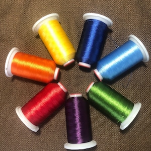 May include: A collection of seven spools of colorful embroidery thread arranged in a circular pattern. The threads are in various vibrant colors, including yellow, orange, red, purple, green, blue, and light blue. Each spool has a white base and top.