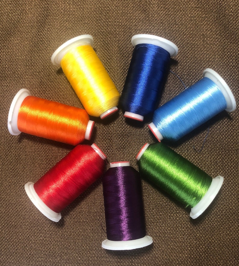 Puede incluir: Una colecci&oacute;n de siete carretes de hilo de bordar de colores brillantes, dispuestos en un patr&oacute;n circular. Los hilos son de varios colores vibrantes, incluyendo amarillo, naranja, rojo, morado, verde, azul y azul claro. Cada carrete tiene una base y tapa blancas.