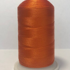 Puede incluir: Una bobina de hilo naranja, ideal para proyectos de costura y manualidades. El hilo est&aacute; enrollado sobre una base de pl&aacute;stico blanco con una parte superior roja. El hilo tiene una textura brillante y suave, perfecto para varios tipos de tejidos.