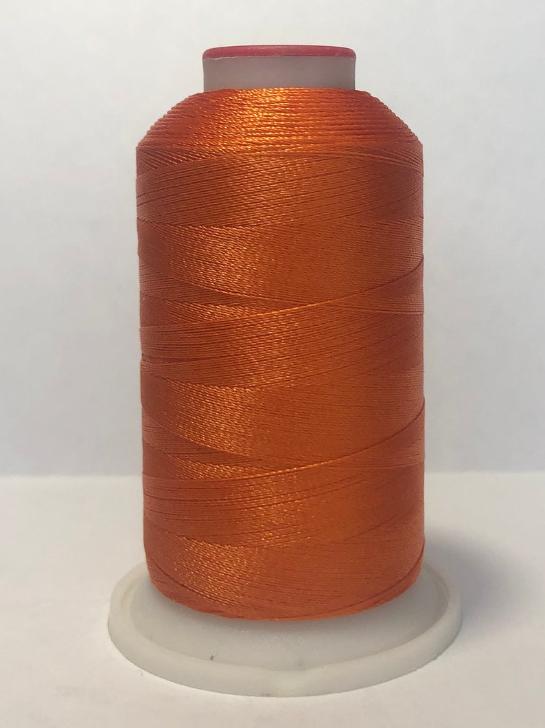 Puede incluir: Una bobina de hilo naranja, ideal para proyectos de costura y manualidades. El hilo est&aacute; enrollado sobre una base de pl&aacute;stico blanco con una parte superior roja. El hilo tiene una textura brillante y suave, perfecto para varios tipos de tejidos.