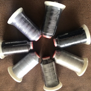 May include: A collection of six spools of thread in various shades of gray and black. Each spool is wrapped in clear plastic and has a white base with a red ring. The spools are arranged in a circular pattern on a brown surface.