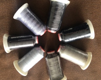 May include: A collection of six spools of thread in various shades of gray and black. Each spool is wrapped in clear plastic and has a white base with a red ring. The spools are arranged in a circular pattern on a brown surface.