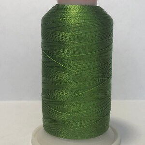 Puede incluir: Una bobina de hilo verde oliva, bien enrollada y brillante, se asienta sobre una base de pl&aacute;stico blanco. El hilo tiene un ligero brillo, lo que sugiere un material de alta calidad. La parte superior de la bobina es de color naranja claro.