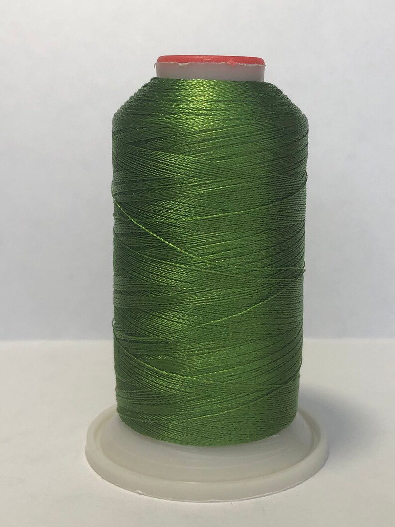 Puede incluir: Una bobina de hilo verde oliva, bien enrollada y brillante, se asienta sobre una base de pl&aacute;stico blanco. El hilo tiene un ligero brillo, lo que sugiere un material de alta calidad. La parte superior de la bobina es de color naranja claro.